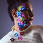 Portrait of a woman with colorful social media stickers on her face, wearing a white sweater.